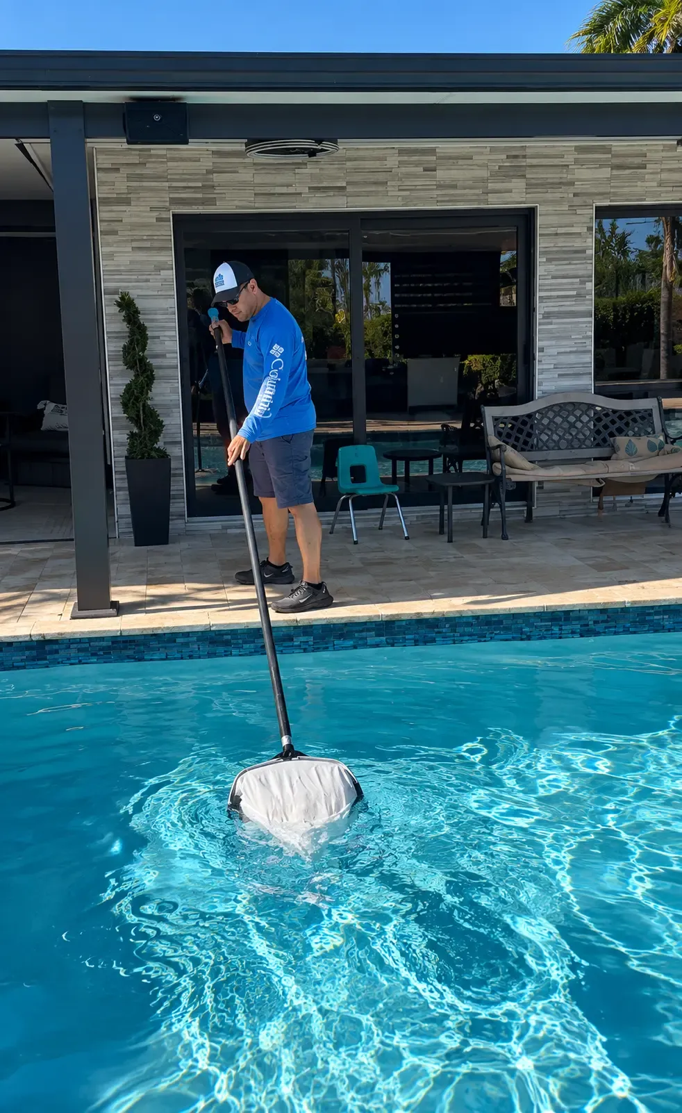 Technician clearing a pool with a leaf net beside a modern tiled home