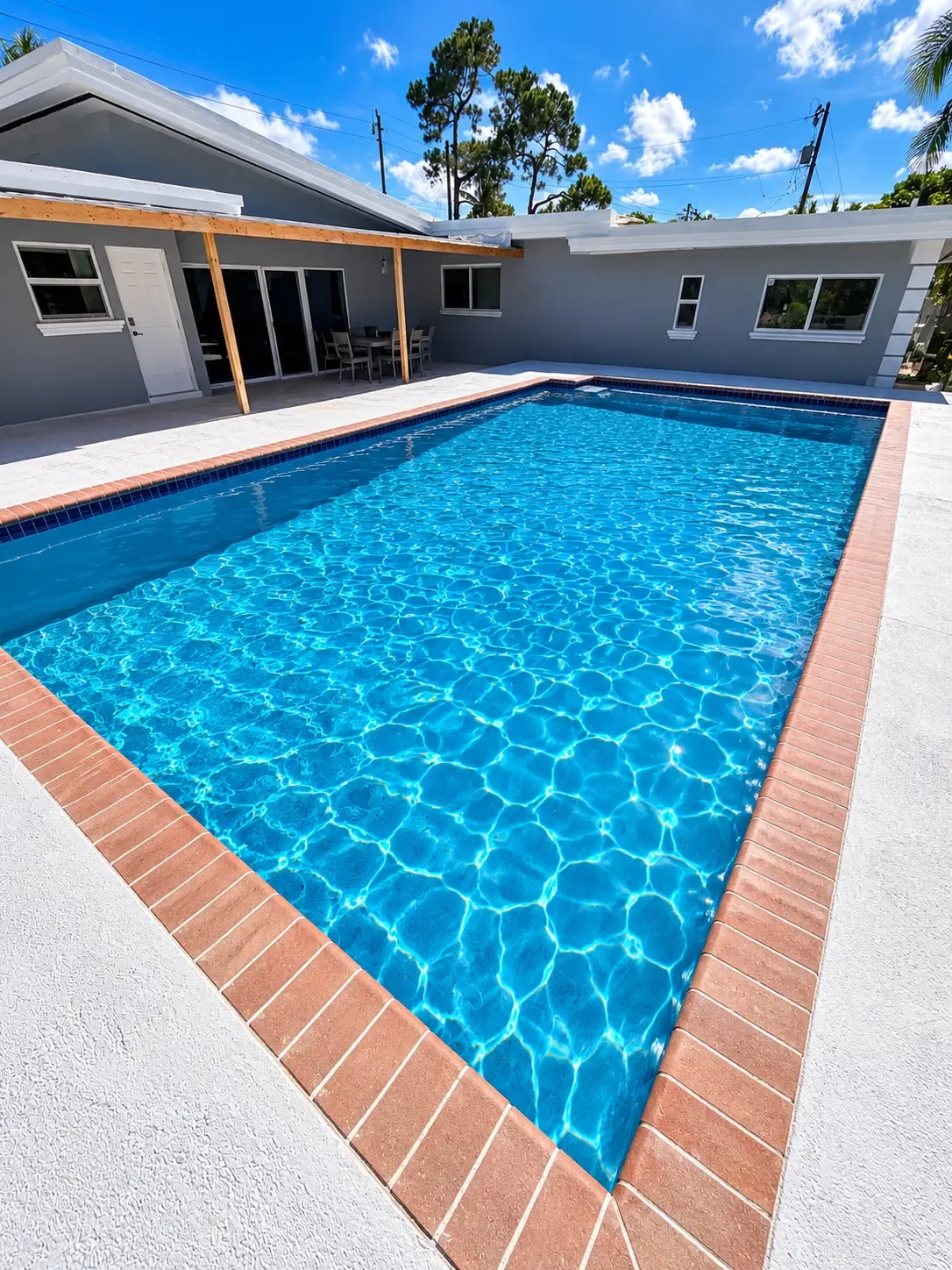 Rectangular pool with brick-red coping and bright blue water behind a gray single-story home